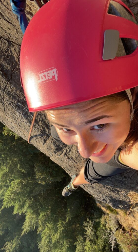 Me sitting on a ledge at the top of the climb. I am wearing a pink helmet and black pants. There is a large drop below me with lots of trees